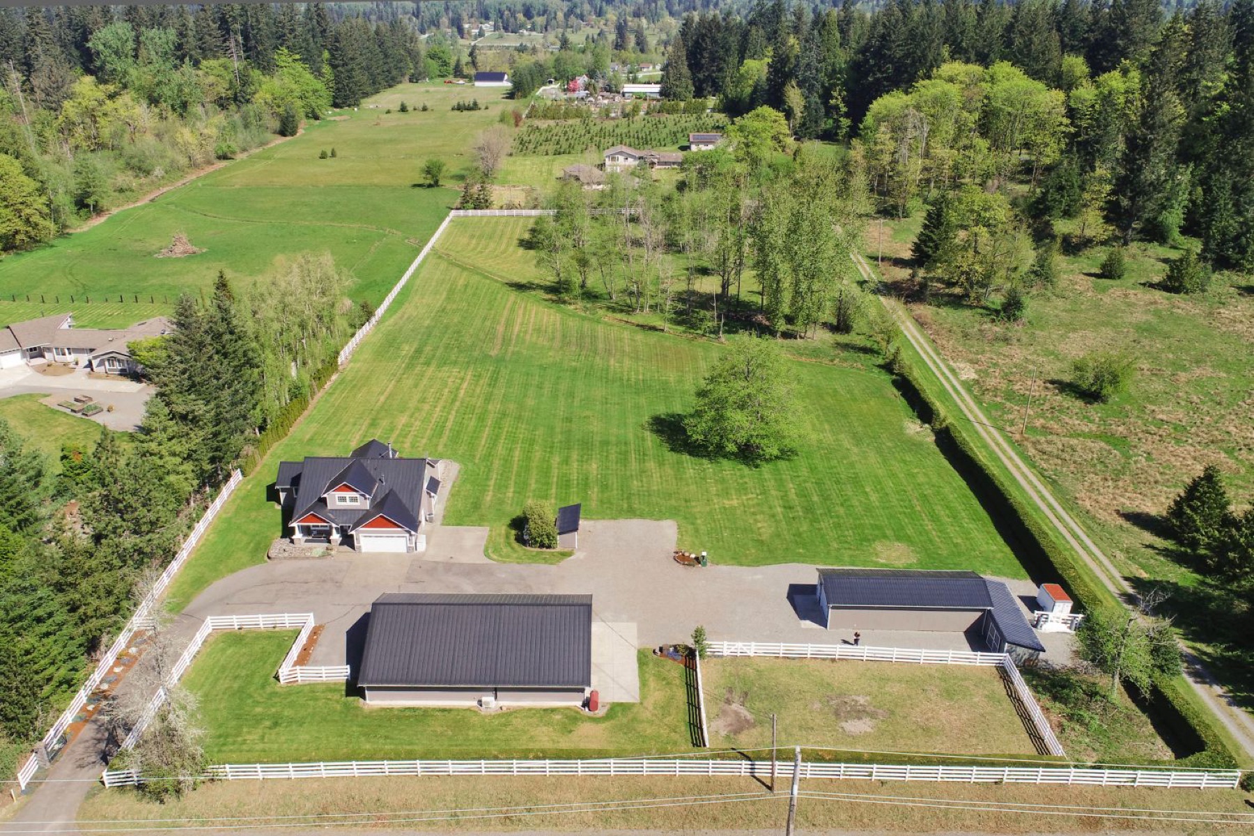 Aerial view of a fenced acreage property with a farmhouse and two shops, surrounded by fields and evergreens